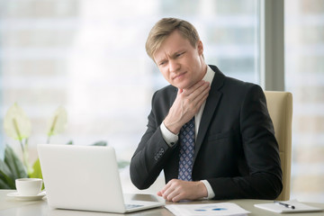 Young businessman working with laptop at desk in the office, hand at his neck, feeling unwell, have a sore throat, after loud screaming, loss of voice, irritation, pain, and itchiness, hard to swallow