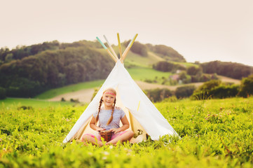 Cute little girl having fun outdoors, playing with tambour, sitting next to teepee © annanahabed