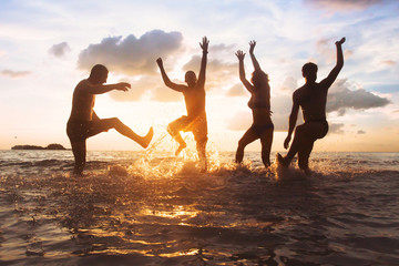group of happy friends having fun together on the beach at sunset, jumping and dancing with water...