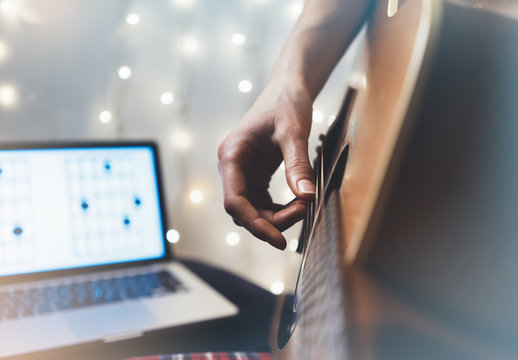Hipster playing guitar in home atmosphere, person studying on musical instrument, notes in laptop on background glow bokeh Christmas illimination, female hands in holiday on relax glitter decoration