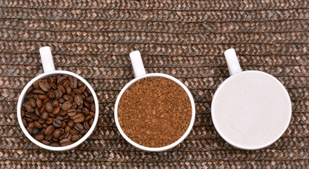 Flat lay. Three white coffee cups with instant coffee, beans and sugar on wicker brown background. Sweet coffee company. Cup of coffee beans, cup of instant coffee an cup of white sugar.