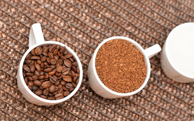 Flat lay. Three white coffee cups with instant coffee, beans and sugar on wicker brown background. Sweet coffee company. Cup of coffee beans, cup of instant coffee an cup of white sugar.