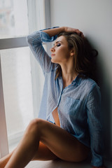 Young pretty woman sitting at window and looking outside enjoys of rest, wearing blue shirt. Studio retouched shot.