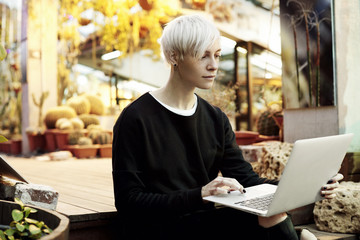 Young hipster woman with blonde short hair sitting on stairs, working on laptop