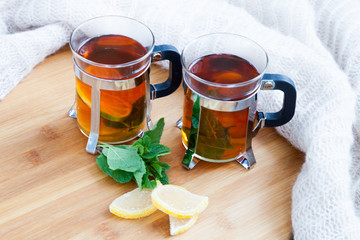 Two glasses of hot black herbal tea with lime, lemon jellybeans, mint leaves on wooden chopping board on table. Light beige knitted scarf on background. Autumn, winter and christmas concept.
