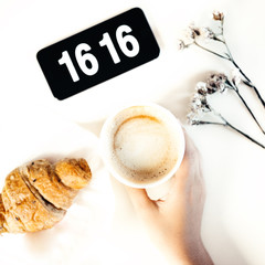 Top view to a composition with white background: croissant, a cup of coffee which is held with a female hand, some dry flowers and a digital clock.