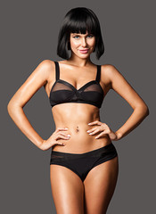 Fototapeta premium Smiling stylish woman in black underwear with a slim figure and tanned body is standing on a gray background