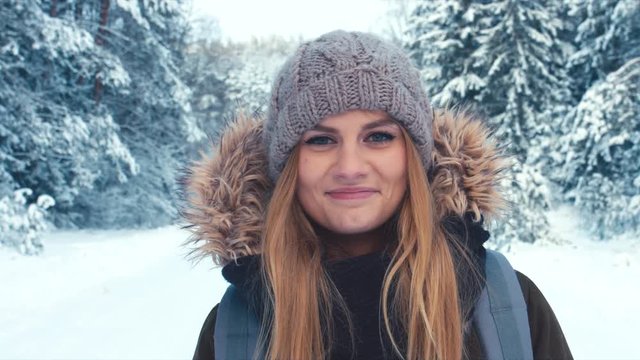 Young attractive Caucasian female hiker with blond hair smiling and looking into the camera, trees covered with snow in the background. Winter scenery, active lifestyle. 4K UHD, 60 FPS SLO MO