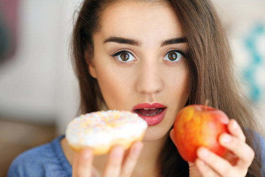 Beautiful young woman making choice between apple and donut, close up