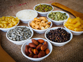 Almond, black pepper seed, Sun flower seed, Cashew nuts, Pumpkin seeds, green bean, rice, fettucine, all put on small white cup and on brown hemp sack texture background