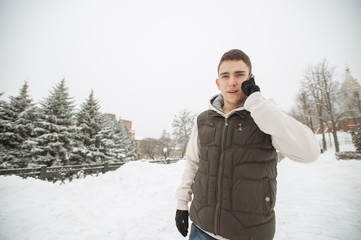 Outdoor winter portrait for young handsome man with the phone. Beautiful teenager in his jacket and vest posing on a city street, background of fir trees.