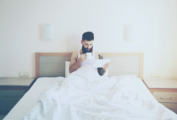 Fototapeta premium Bearded man lying in morning bed with coffee or tea cup and digi