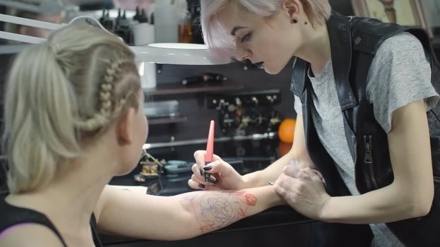 Young woman drawing flower tattoo design on skin of arm of her female client in studio