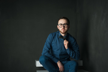 studio portrait of a young man on a dark wall background in a blue shirt and jeans. hipster man, dressed stylishly, with a beard and glasses, sitting on white stairs