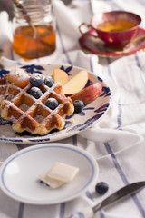Blueberry apple waffle with honey set on a dish decorated with fresh blueberry, apple, tea pot set ready to serve.