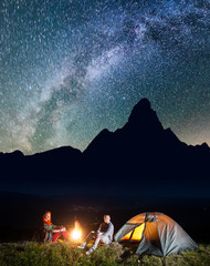 Happy couple hikers sitting near campfire and lighting tent under incredibly beautiful starry sky. In the background silhouette of the mountains and luminous village in the valley at night. Low light