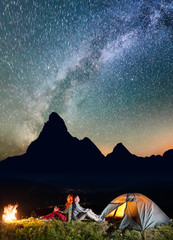 Romantic couple backpackers sitting by bonfire near tent under incredibly beautiful starry sky. Silhouette of the mountains and luminous village in the valley at night in background. Astrophotography