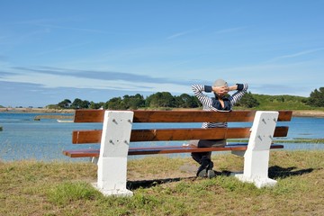 Une jeune femme habillée d'un pull marin est assise sur un banc face à la mer