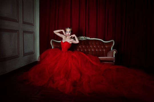 Beautiful girl in long red dress and in royal crown sitting on the retro sofa  on red background in photo studio