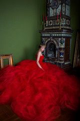 Beautiful girl in long red dress and in royal crown near fireplace in luxury interior in photo studio