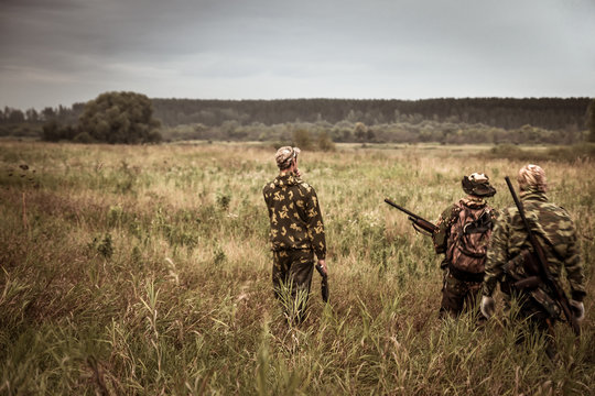 Hunters in camouflage walking through rural field during hunting season season in overcast day with moody sky