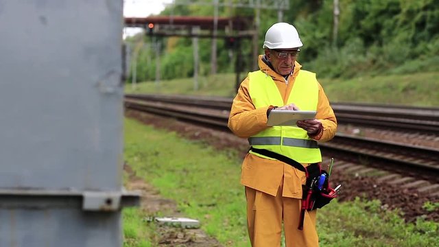 Railwayman makes notes in his tablet computer. Railway employee in yellow uniform on railway line. Railway worker in yellow uniform and white hard hat with tablet pc in hands