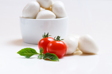 Small mozzarella balls in white bowl with cherry tomatoes and fresh basil on white background
