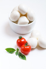 Small mozzarella balls in white bowl with cherry tomatoes and fresh basil on white background