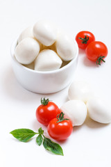 Small mozzarella balls in white bowl with cherry tomatoes and fresh basil on white background