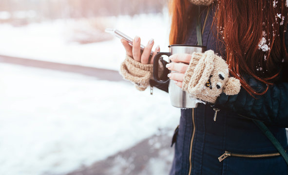 Winter. Woman with red hair wearing gloves . Girl drinking hot tea or coffee  iron insulated cup. It uses a phone  smartphone. Playing, looking for something on the Internet. Scrolls news feed