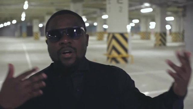Dolly shot of African man in suit and sunglasses walking in parking lot and rapping and gesturing