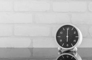 Closeup black and white alarm clock for decorate in 6 o'clock on black glass table and white brick wall textured background in black and white tone with copy space