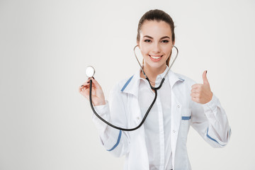 Cheerful young woman doctor using stethoscope and showing thumbs up