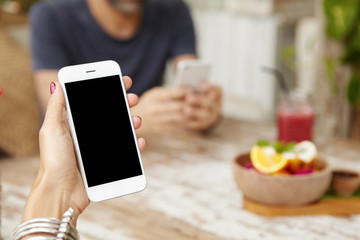 Close up view of woman's hands with red nails wearing stylish bracelet holding mobile phone, showing something on its screen while relaxing at cafeteria during lunch with her boyfriend in background