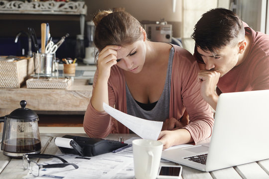 Indoor shot of unhappy young family distressed with financial problems and mounting bills, reading document with frustrated looks while calculating domestic finances together in their kitchen