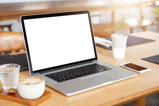 Mockup of generic laptop computer with blank copy space screen for your text or advertising content. Workspace of young freelancer: open notebook pc, cell phone and cappuccino on wooden table