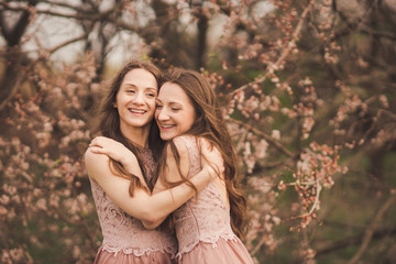 Beautiful happy young twins sisters in long evening dresses enjoying smell in a blooming spring garden. Having fun together, positive emotions, pastel colors. Healthy hair and clean skin. Copy space.
