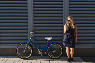 Fototapeta na wymiar Lifestyle and technology. Young pretty blonde woman using her smartphone standing with her vintage bike on urban wall background. Good day for a ride. Copy space.