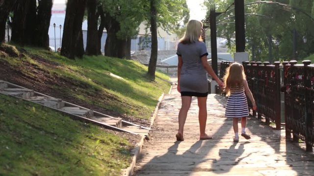 Pregnant mother and her little happy daughter walk in sunny park, back view