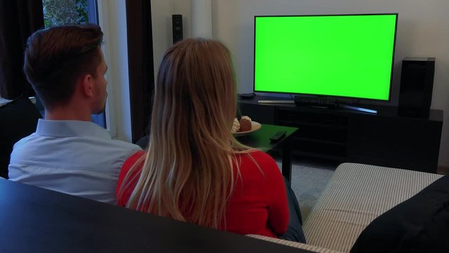 A young couple watches a TV with a green screen in a cozy living room
