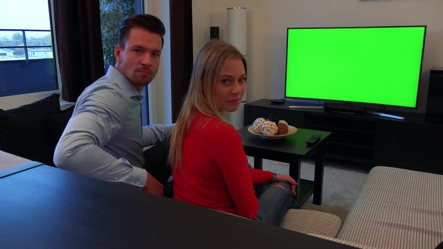A man and a woman watch a TV with a green screen in a cozy living room, then turn to the camera and shake their heads dissatisfiedlyv