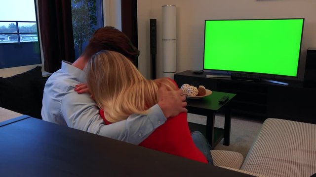 A young couple in an embrace watches a TV with a green screen in a cozy living room and cries