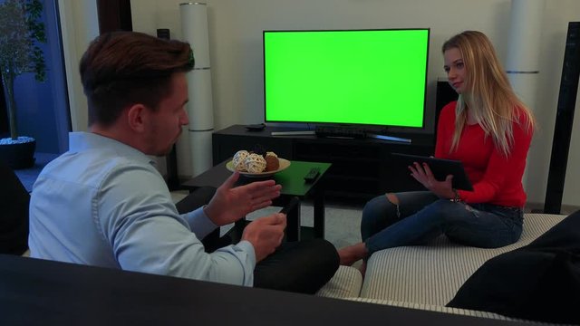 A young, attractive couple on a couch in a cozy living room work on a smartphone and a tablet, the man explains something to the woman - TV with a green screen in the background