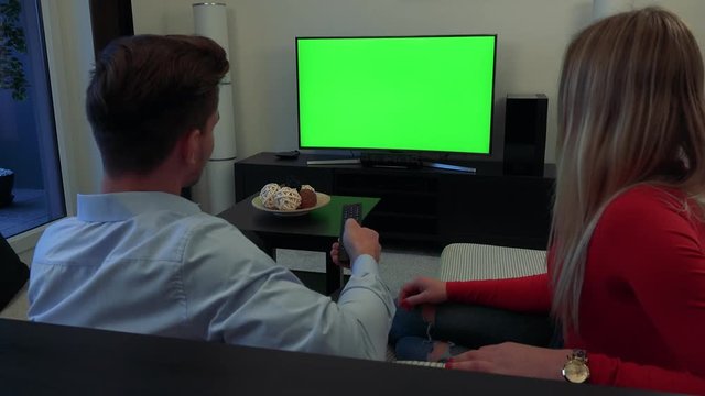 A young, attractive couple watches a TV with a green screen in a living room, the man switches between channels, the woman shakes her head and takes over the controller