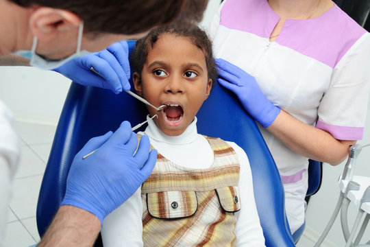 little African-American ethnic black female smiling while dentist in white latex gloves check condition of her teeth. baby girl in blue dental chair