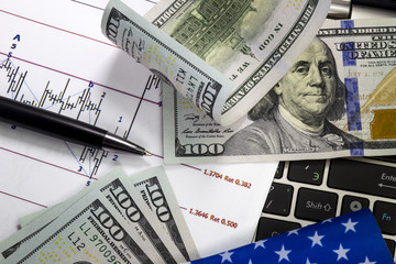 Stock market prices chart. Angled hundred us dollar bills above the chart.Rolled bill lying on another bill. Notebook keyboard under the graph. Pen pointing on the graph. Us flag.Macro image.
