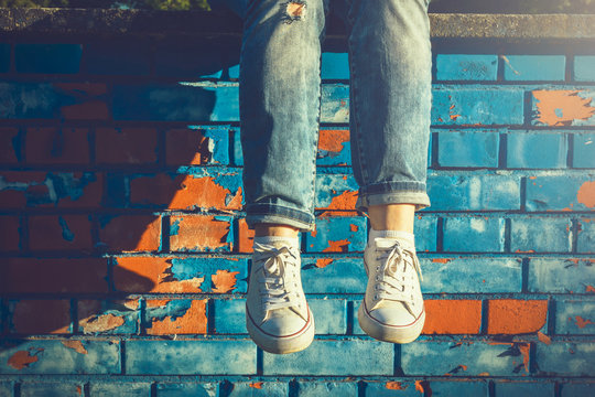 Female legs in white canvas sneakers and jeans dangling from a blue brick wall on a sunny summer day