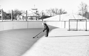 Four year old toddler girl on skates at an outdoor ice skating rink learning to play hockey with stick and puck in black and white