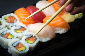 Hand using chopsticks pick Sushi and Sashimi rolls on a black stone slatter. Fresh made Sushi set with salmon, prawns, wasabi and ginger. Traditional Japanese cuisine..