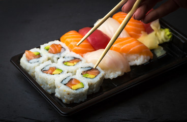 Hand using chopsticks pick Sushi and Sashimi rolls on a black stone slatter. Fresh made Sushi set with salmon, prawns, wasabi and ginger. Traditional Japanese cuisine..
